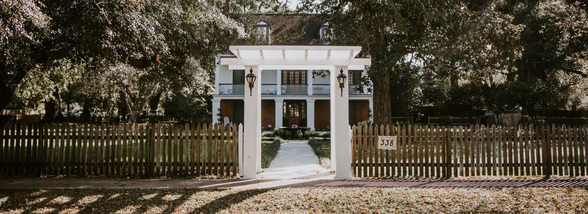A white house with a dark roof and porch surrounded by trees and a wooden fence.