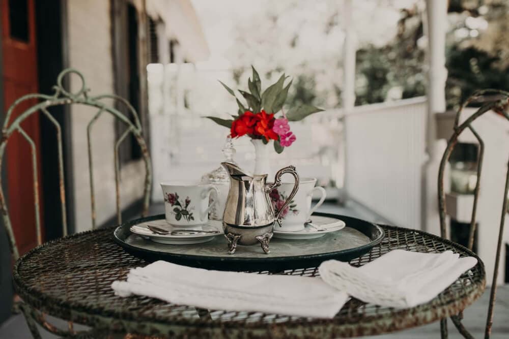 A small outdoor table set with a silver teapot, floral teacups, and a vase of fresh flowers.